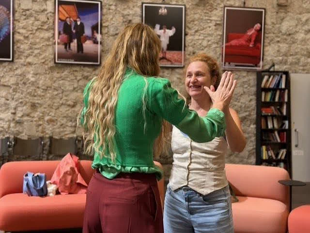 Élodie Tisserand, souriante, travaille face à une comédienne vue de dos, la main levée près du visage, dans une salle aux murs de pierre garnie d'affiches de spectacle.